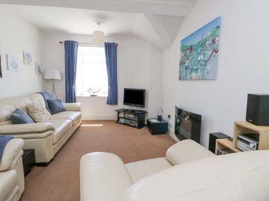 A living room with beige sofas, a small TV on a stand, blue curtains, and a painting on the wall at Bethan Apartment in Trearddur Bay
