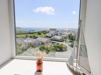 A window view of houses and the sea with a small sailboat model and a jar with dried flowers on a windowsill at Bethan Apartment in Trearddur Bay