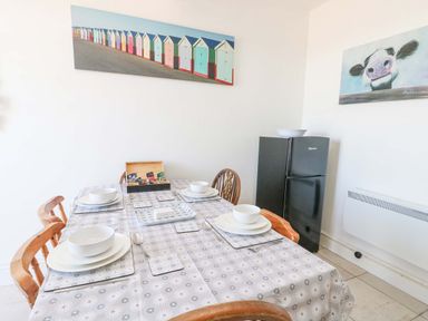 A dining table with four place settings and wooden chairs next to a black refrigerator in a room with wall art at Bethan Apartment Trearddur Bay