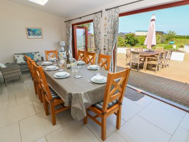 A dining room with a table set for eight and wooden chairs opening to an outdoor patio with a table and umbrella at Llain Gwynfor in St Davids