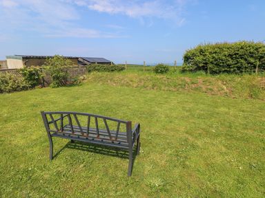A metal bench on a grass lawn with bushes and a building with solar panels in the background at Llain Gwynfor in St Davids