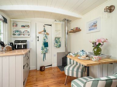 A kitchen area with countertop appliances and dishes hanging on the wall next to a small dining table with striped chairs and a vase of flowers at Collen in Aberaeron