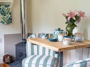 A wooden table with a teapot cups jar and vase with flowers beside striped chairs in a room with a wood stove at Collen in Aberaeron