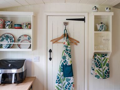 A kitchen door with aprons hanging on wooden hangers a shelf with patterned plates bowls and cups an air fryer on a counter and a small shelf with a teapot glasses and a patterned towel at Collen in Aberaeron