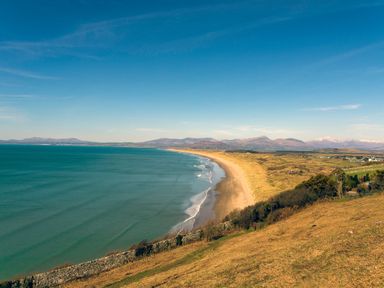 A wide sandy beach with gentle waves beside grassy dunes and hills with mountains in the background at Collen in Aberaeron