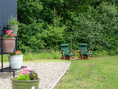 Two green cushioned wooden chairs and a small wooden table on grass near bushes and flower pots on a gravel area at Bedwen in Aberaeron