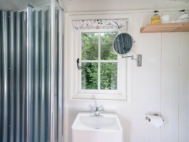 A bathroom with a window above a sink a round wall mirror a metal shower enclosure and a wooden shelf with toiletries at Bedwen in Aberaeron