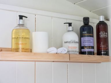 A wooden shelf with bottles of Baylis and Harding toiletries and small rolled white towels on a white paneled wall at Bedwen in Aberaeron