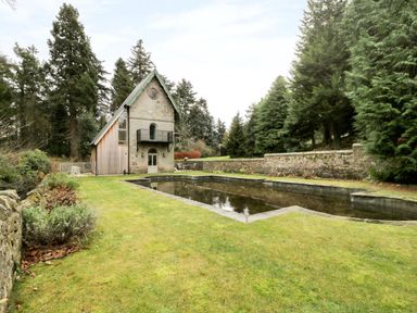 A stone and wood house with a black balcony next to a rectangular reflecting pool surrounded by grass and trees at The Folly in Belford