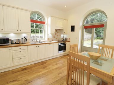 A kitchen with wooden cabinets, countertop appliances, a glass dining table with chairs, and arched windows at The Folly in Belford