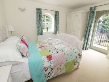 A bedroom with a double bed covered in a patchwork quilt and pillows with a floral pattern next to a window with turquoise floral curtains and a white wardrobe at The Folly in Belford