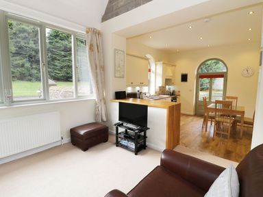 An open living area with a brown leather sofa and ottoman a TV on a stand a dining table with chairs and a kitchen with white cabinets at The Folly in Belford