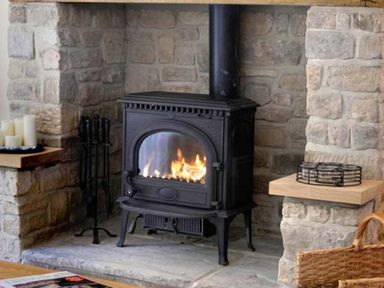 A stone fireplace with a black wood-burning stove fire tools candles and a wicker basket Near Belford