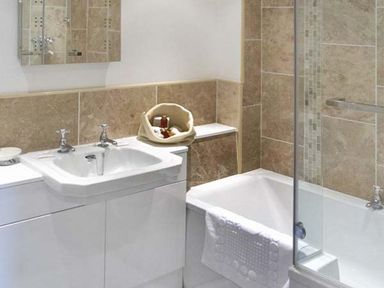 A bathroom with a white sink and cabinet next to a bathtub with a white towel and beige tiled walls at The Lake Cottage Near Belford