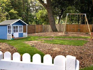 A fenced backyard with a blue playhouse a double swing set and wood chip ground cover near Belford