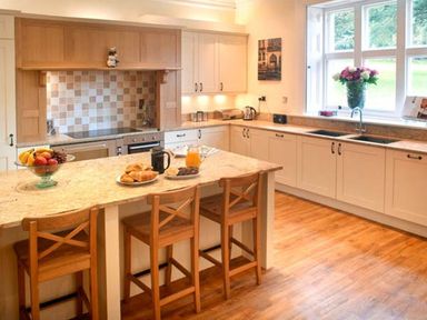 A kitchen with a marble island and wooden stools a fruit bowl flowers in a vase and a window at The Hall in Belford