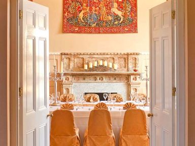 A dining room with a set table and chairs covered in gold fabric in front of a fireplace with candles and a tapestry at The Hall in Belford