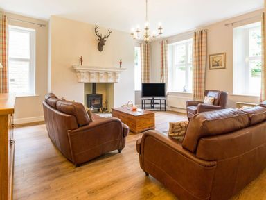 A living room with brown leather sofas a wooden coffee table a TV on a stand a fireplace with a mounted deer head and plaid curtains at The Coach House in Belford