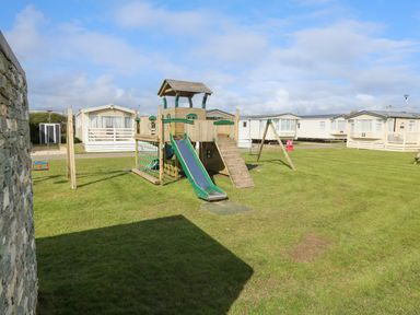 A playground with a slide climbing wall swings on a lawn surrounded by mobile homes at Cornerstone Near Caernarfon