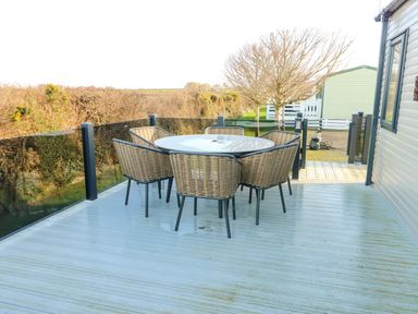 An outdoor deck with a round table and six wicker chairs near a caravan at Cornerstone Near Caernarfon