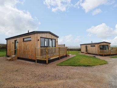 Two wooden cabins with small porches on a gravel and grassy area under a partly cloudy sky at Cae Dafadd in New Mills near Llanfair Caereinion