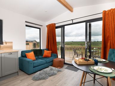A living area with a blue sofa, orange cushions, a round dining table set for two, and sliding glass doors opening to a balcony with countryside views at Cae Dafadd New Mills near Llanfair Caereinion