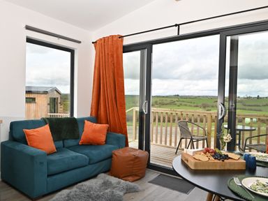 A living room with a blue sofa orange cushions and curtain a brown pouf glass sliding doors leading to a wooden deck with chairs and green fields beyond at Cae Dafadd in New Mills near Llanfair Caereinion