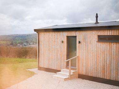 An exterior view of a small wooden cabin with a door and steps overlooking a rural landscape at Cae Bedw in Llanfair Caereinion