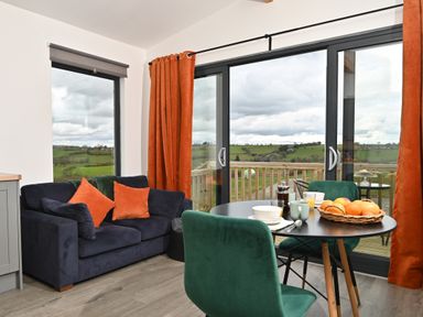 A living area with a dark blue sofa with orange cushions a round dining table with green chairs and glass doors overlooking a green landscape at Cae Bedw in Llanfair Caereinion