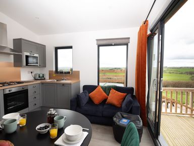 A kitchen and seating area with a round table set with cups and bowls a blue sofa with orange cushions and sliding glass doors opening to a deck with a view at Cae Bedw Llanfair Caereinion