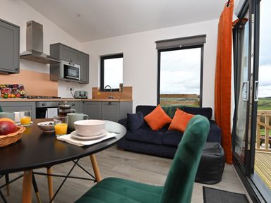 A kitchen and dining area with grey cabinets a black table with breakfast items and a navy sofa with orange cushions at Cae Bedw Llanfair Caereinion