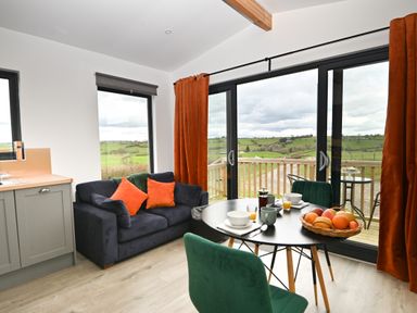 A small living area with a navy sofa with orange cushions next to a dining table set for breakfast and sliding glass doors overlooking green fields at Cae Bedw in Llanfair Caereinion