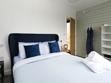 A bedroom with a bed with blue and white bedding and pillows two rolled white towels on the bed a wooden door a side table with a lamp and tray two dark robes hanging on a white shelving unit at Cae Bedw in Llanfair Caereinion