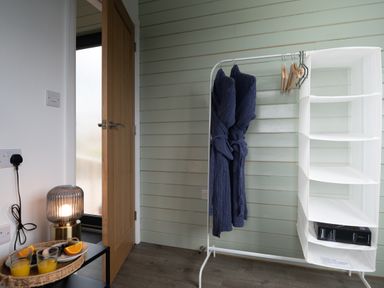 A small area with a wooden door open showing a frosted glass window two robes hanging on a white clothing rack and a table with a lamp and a tray of drinks and fruit at Cae Bedw in Llanfair Caereinion
