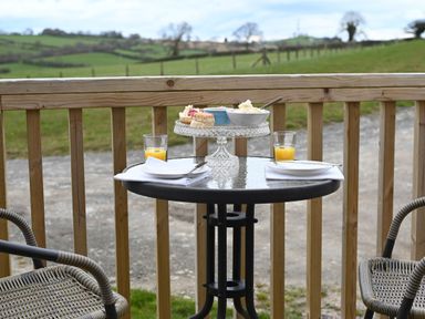 An outdoor table with two glasses of orange juice two plates with cutlery a cake stand with scones and bowls of cream on a wooden deck overlooking green fields at Cae Bedw in Llanfair Caereinion