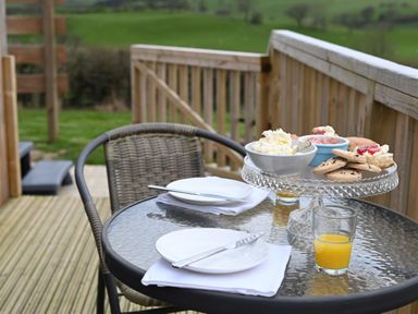 A glass outdoor table with plates knives napkins a glass of orange juice and a glass cake stand with cream jam scones and cookies on a wooden deck with a wicker chair at Cae Bedw Llanfair Caereinion