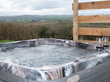 An outdoor hot tub with bubbling water and two glasses of red wine near a wooden fence with green hills in the background at Cae Bedw in Llanfair Caereinion