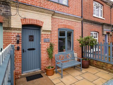 Front door and window of a brick cottage with a blue bench and potted plants at Compass Cottage in Aldeburgh