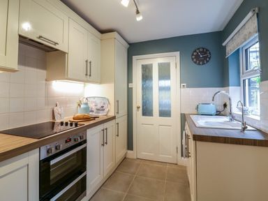 A kitchen with white cabinets a countertop with bread a kettle a toaster a clock on the wall and a sink by a window at Compass Cottage in Aldeburgh