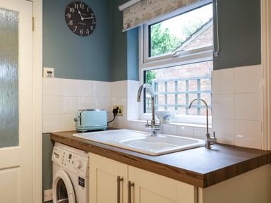 A kitchen counter with a washing machine a double sink a blue toaster and a wall clock at Compass Cottage in Aldeburgh
