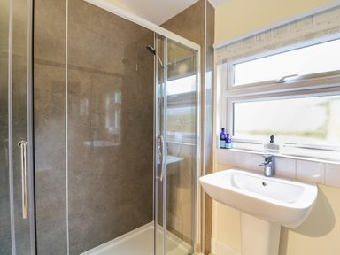 A bathroom with a glass shower enclosure and a white pedestal sink under a window at Compass Cottage in Aldeburgh