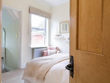 A bedroom with a bed covered by a textured blanket a window with a blind a pink armchair and a wall with shelves holding a model sailboat and a mirror at Compass Cottage in Aldeburgh