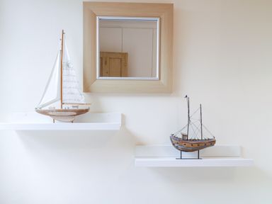 Two model sailboats on white wall shelves beneath a square wooden framed mirror at Compass Cottage in Aldeburgh