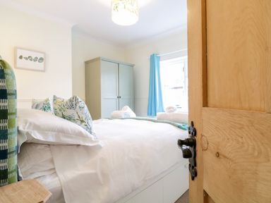A bedroom with a bed pillows a wooden door a wardrobe and a window with blue curtains at Compass Cottage in Aldeburgh
