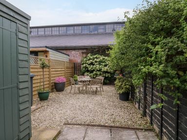 A gravel patio area with a metal table and four chairs surrounded by potted plants and wooden fences at Compass Cottage in Aldeburgh