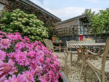 An outdoor seating area with metal chairs and table next to pink flowers and shrubs at Compass Cottage in Aldeburgh
