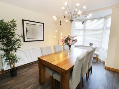 A dining room with a wooden table and six upholstered chairs a vase with flowers on the table a large window with blinds a potted plant and a framed picture on the wall at Old Post Office in Blaenau Ffestiniog