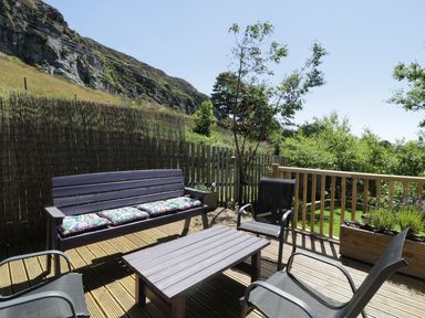An outdoor deck with a wooden bench, cushions, chairs and a table with a view of a hill and trees at Old Post Office in Blaenau Ffestiniog