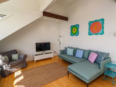 A living room with a green sectional sofa with cushions a gray armchair a white TV stand with a television two colorful paintings on the wall and a brown rug on the floor at Ocean View in Alnmouth