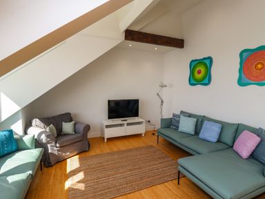 A living room with a gray armchair a teal sofa a white TV stand with a television and colorful wall art at Ocean View in Alnmouth